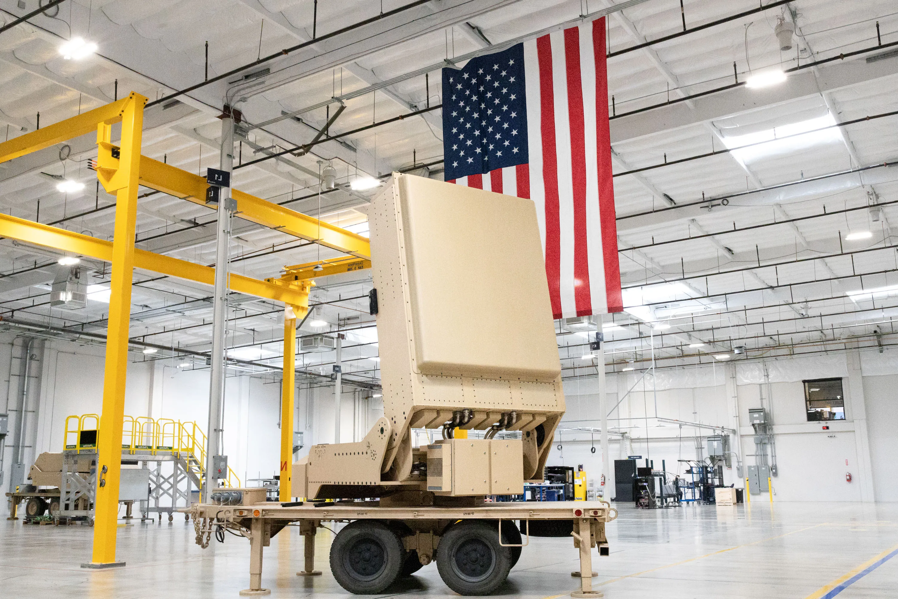 A large tan piece of military equipment sits on a trailer in a warehouse with a large American flag hanging from the ceiling.